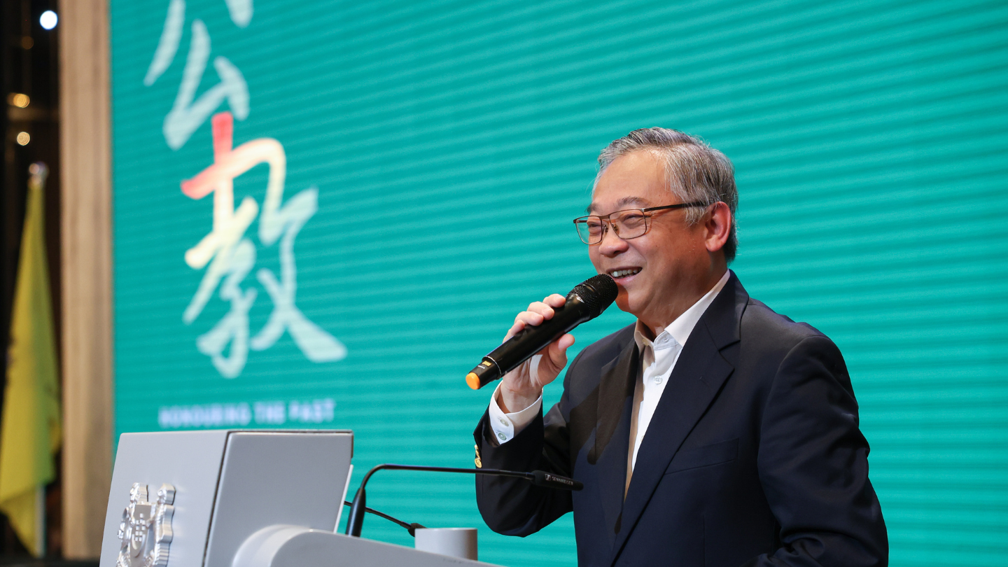 Smiling speaker with microphone at podium; teal backdrop with large Chinese characters.